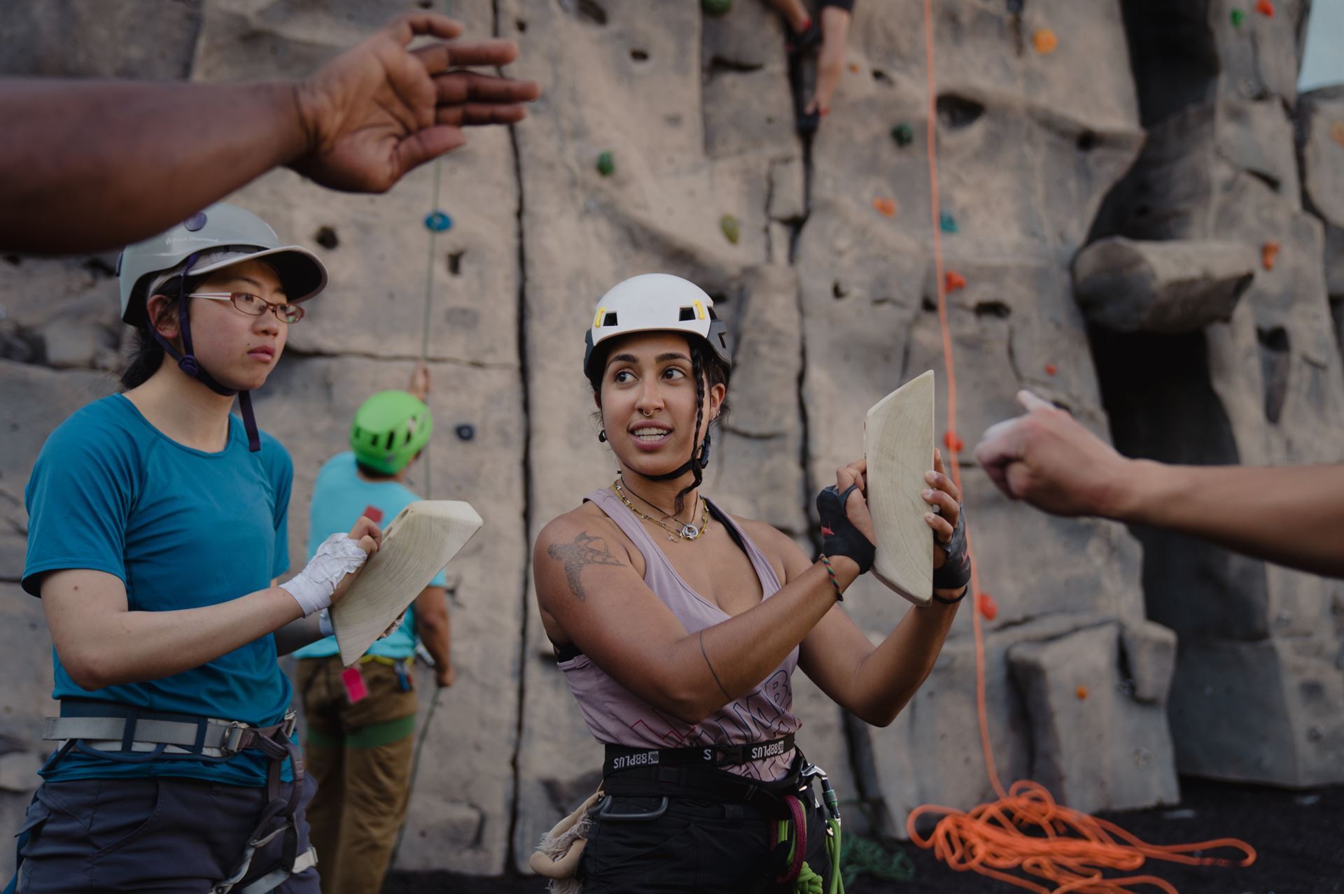 Sof stands in a climbing gym in a climbing harness and helmet. She is demonstrating grip type on a wooden block for a group of climbers.