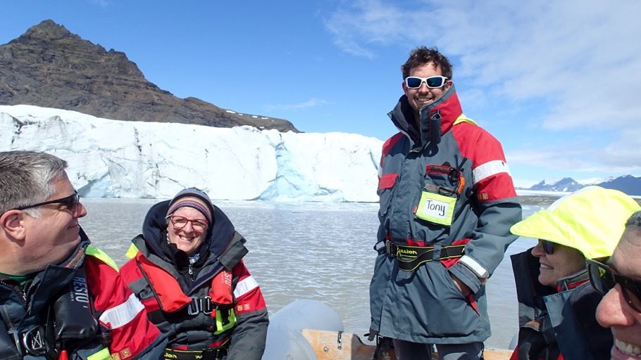 Tony stands next to a group of people on a boat. There is water and ice in the background. Tony and the group wear red and grey jackets.