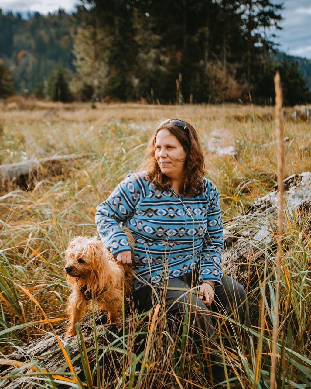 White femme non-binary person sitting on log beside their small dog