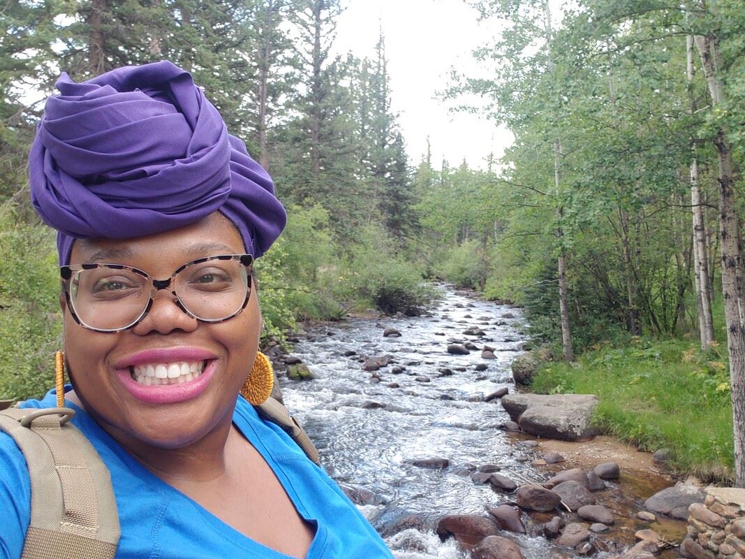 A selfie of Parker standing by a creek in the forest. They are wearing glasses, a blue shirt, and yellow earrings.